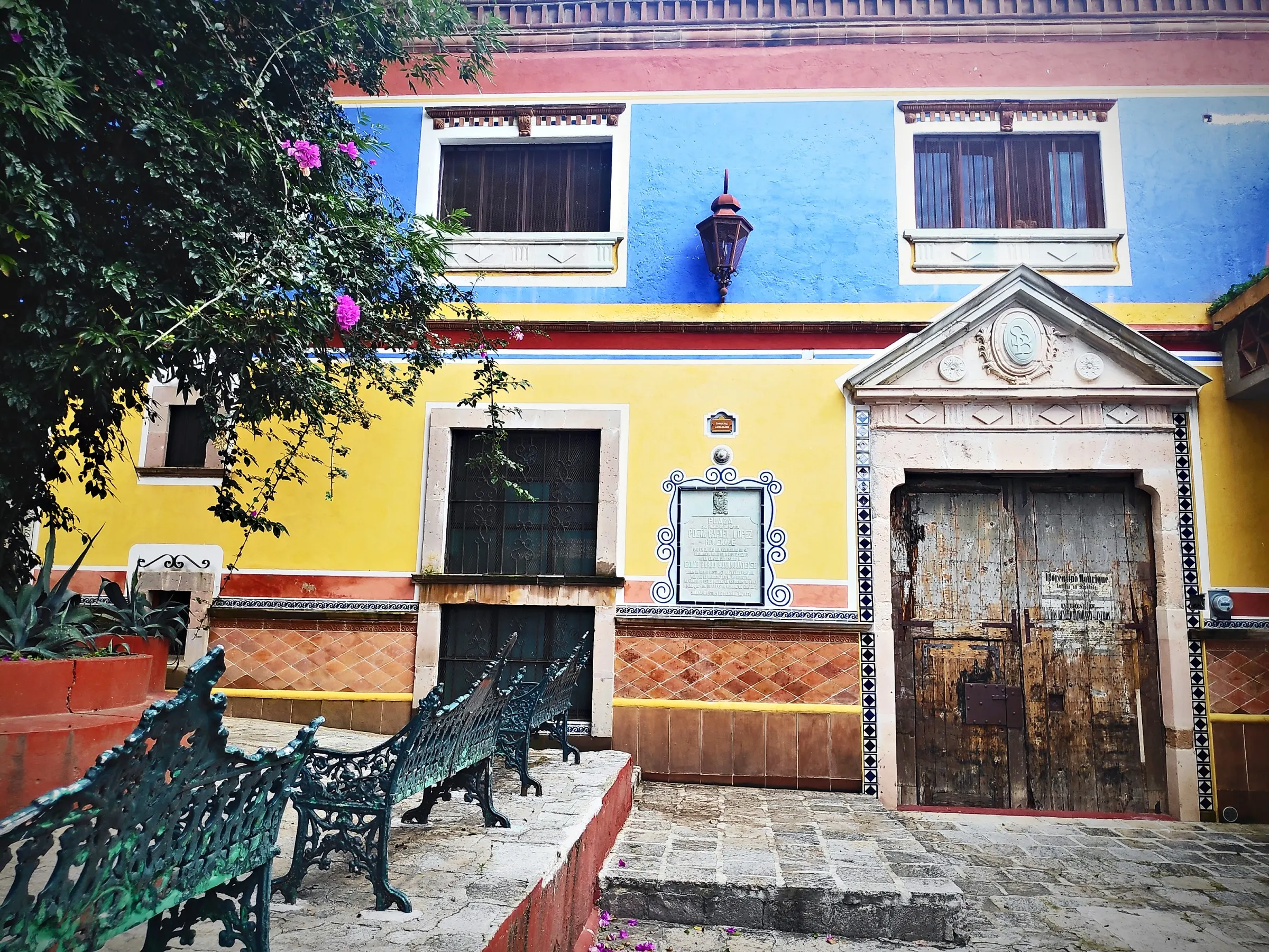 Blue and yellow façade with a carved stone doorway and iron benches on the little square