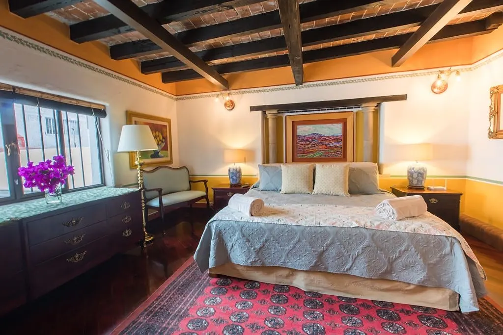 Los Picachos suite: bed beneath a wooden-beam ceiling, with a landscape painting and flowers at the window