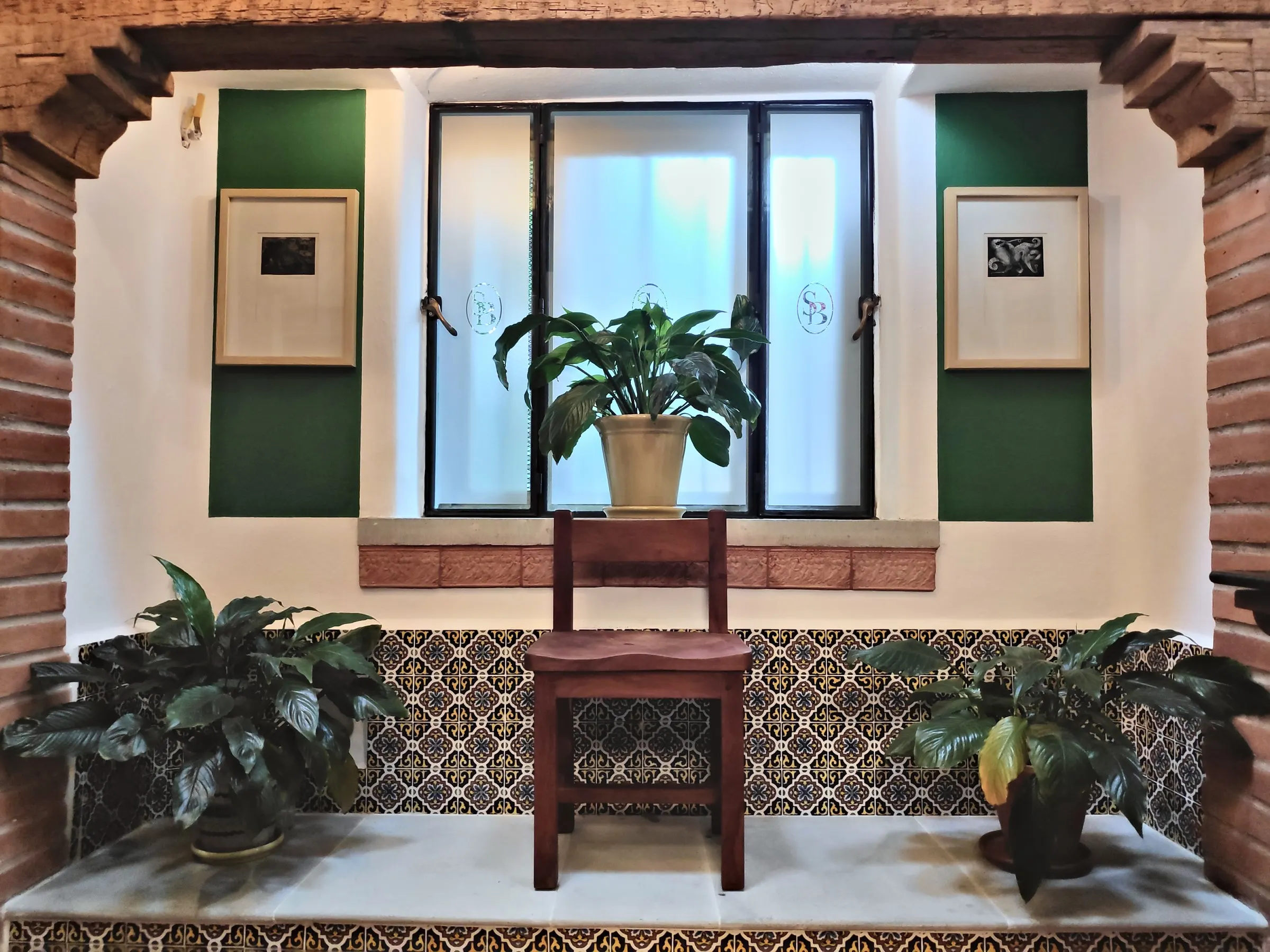 Window between green walls with a wooden chair, plants and a Talavera bench